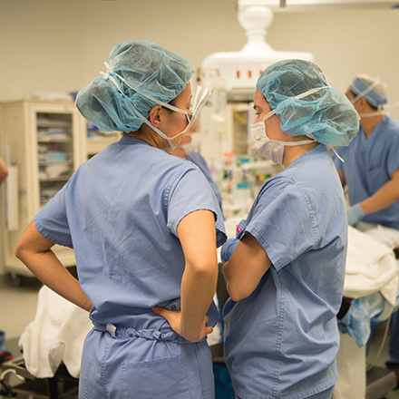 Two female providers wearing surgical caps and masks have a discussion in a trauma room